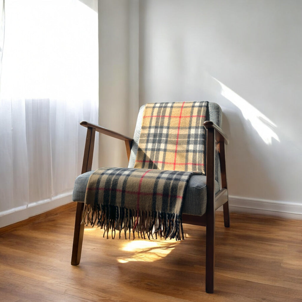 A  camel thomson recycled wool tartan knee blanket with fringed edges draped over a wooden chair in a room with a light interior.
