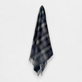 A dark navy and gray plaid scarf with fringed edges, hanging against a plain light gray background.