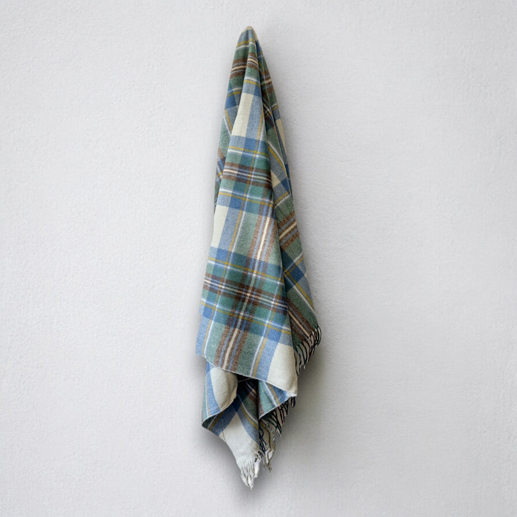 a 100% recycled blanket in stewart muted blue tartan hanging from a wall hook on a white background 